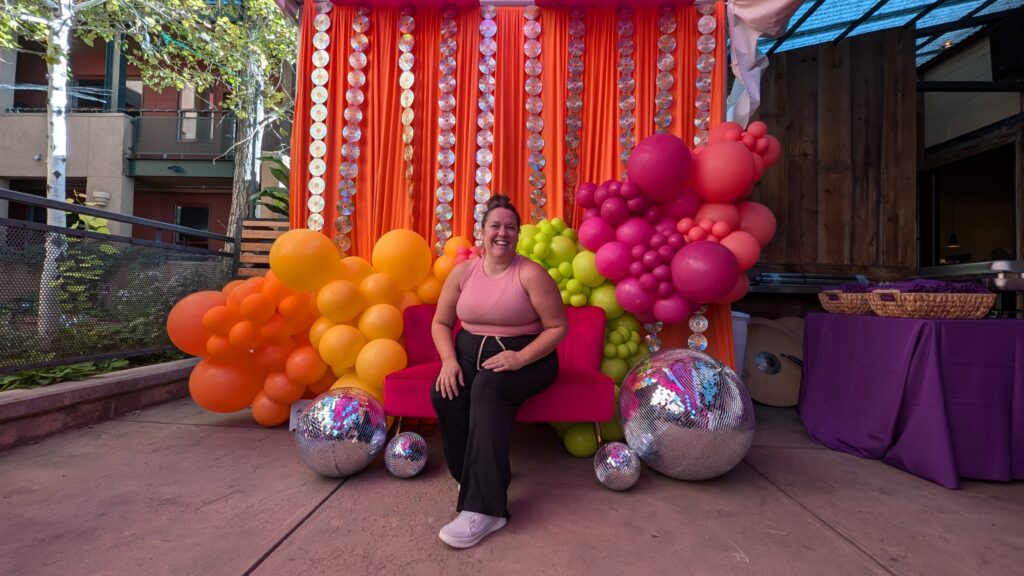 white woman with brown hair named Missy Castaneda from Glenwood Springs, Colorado is the owner of The Fluffy Stuff balloon decor and event styling. She is seated on a bright pink sofa with disco balls and colorful balloons around her and CDs hang behind her on an orange background